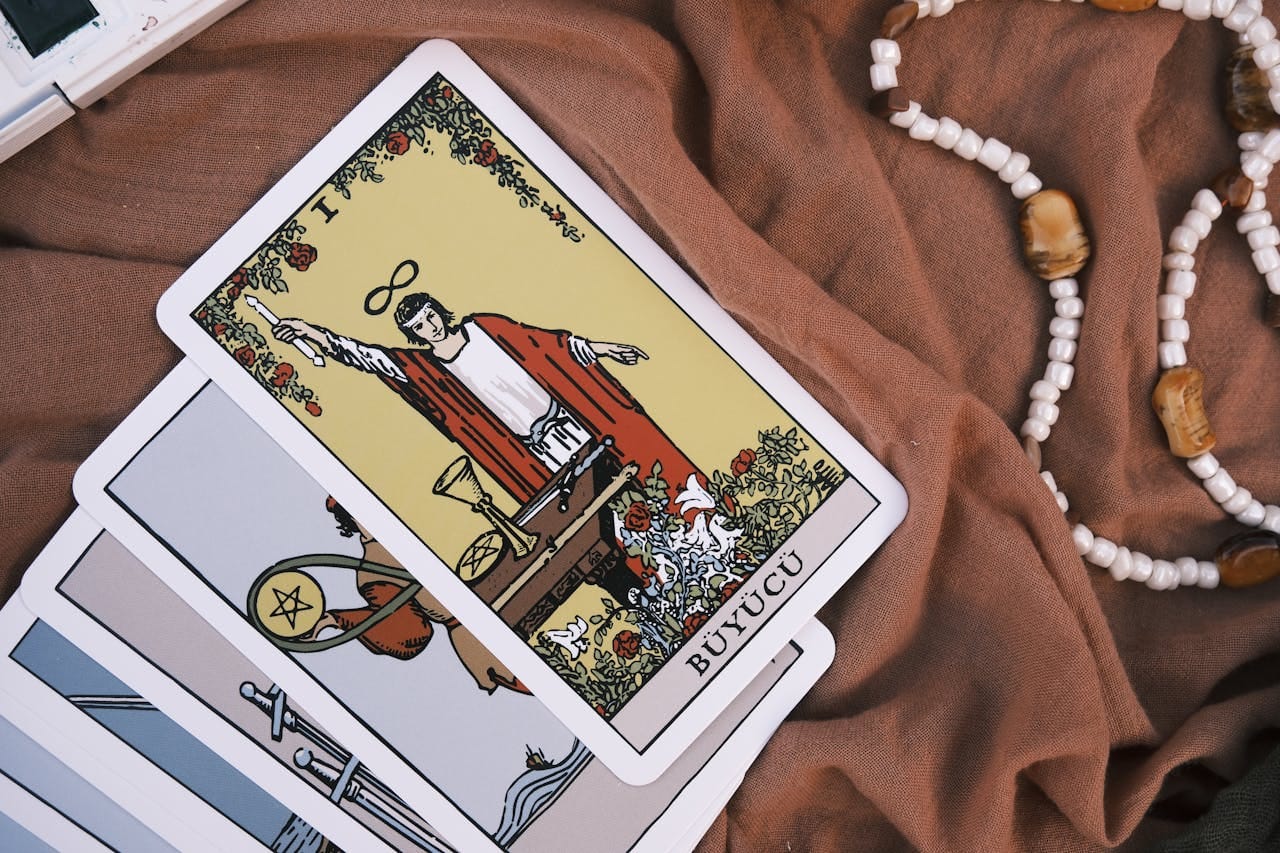 A close-up of tarot cards with a beaded necklace on a fabric surface, featuring the Magician card.